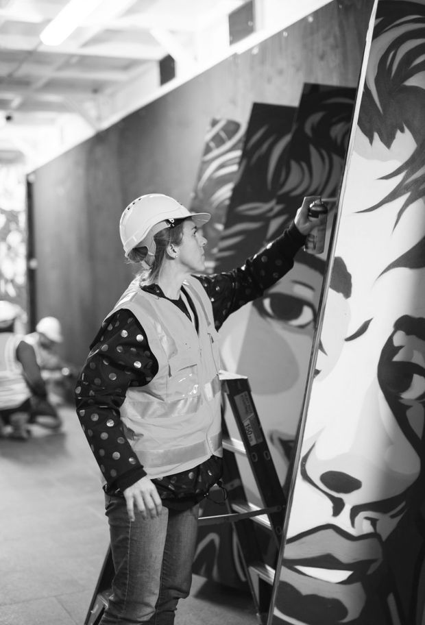 Black-and-white photograph of a worker in a hard hat and high-visibility vest installing or working on a large graphic mural panel in an indoor public or commercial space. A ladder, unfinished wall surfaces, overhead lighting, and large monochrome portrait artwork are visible. The image focuses on the installation process rather than a finished room.