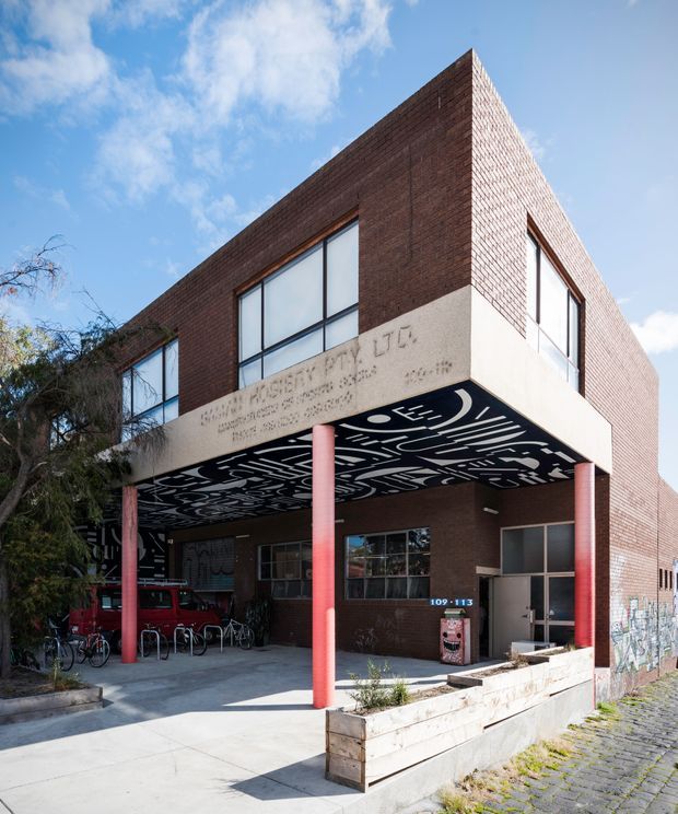 Exterior architectural photograph of a multi-storey institutional or commercial brick building with a prominent projecting canopy, large windows, red cylindrical support columns, and a black-and-white graphic soffit. A paved entrance forecourt, bicycle racks, planter boxes, and signage are visible.