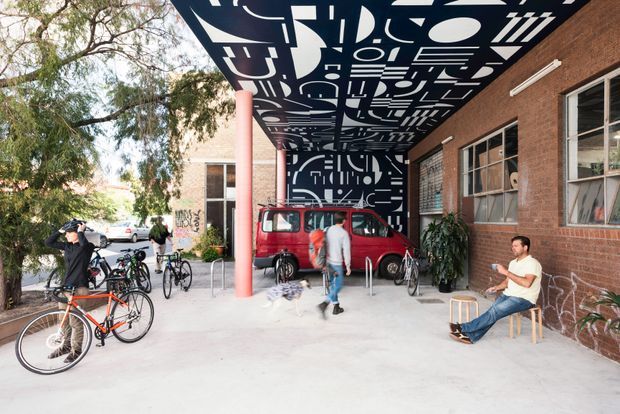 An open-air covered entrance or courtyard beside a brick building, with large industrial-style windows, a striking black-and-white geometric mural across the ceiling and rear wall, a bright red vintage van, bicycles, and casual seating. The space has a creative urban commercial or community setting with exposed brick, painted steel columns, and concrete paving.