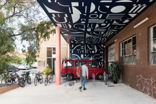 Exterior view of a brick building with a covered pedestrian forecourt or entry canopy. The canopy soffit and rear wall feature bold black-and-white geometric graphic artwork, supported by a bright coral-red column. A red van, bicycles, people, and planting occupy the forecourt, while mature trees and neighbouring buildings are visible beyond.