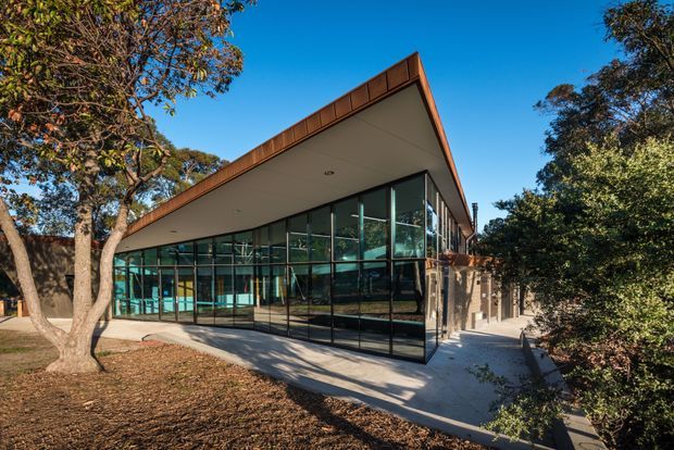 Exterior view of a low-profile modern building set among mature trees, with extensive floor-to-ceiling glazed curtain walls, a projecting flat roof with warm timber-toned fascia, and a paved entrance walkway. The transparent facade reveals an open interior but the building exterior dominates the image.