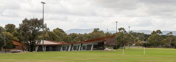 Low, single-storey contemporary sports or community pavilion set beside a broad grass field, with a long dark glazed facade, angled structural elements, and a shallow reddish-brown roof. Tall sports floodlights, goal posts, mature trees, and distant hills are visible under an overcast sky.