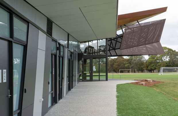 Exterior view of a contemporary sports or community facility beside a grass soccer field. The building has dark metal-framed glazing, grey panel cladding, a broad flat soffit, and a prominent angular perforated metal canopy or sunshade projecting over the entrance walkway. The covered path connects glazed rooms to the playing fields.