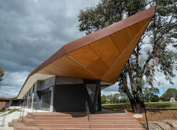 Exterior view of a contemporary, angular building with a dramatic projecting canopy clad in warm timber panels and edged with rust-red metal. The facade combines dark panels, glazing, and light wall surfaces, accessed by broad steps with metal handrails. The building sits beside landscaped grounds and mature trees under a cloudy sky.