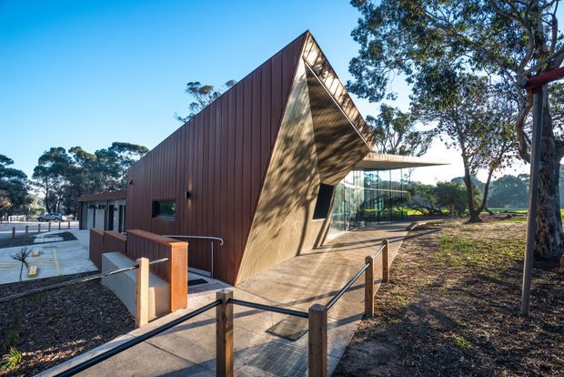 Exterior view of a contemporary single-storey building set among mature trees. The building has a dramatic steeply pitched, angular gable form clad in vertically ribbed dark timber or metal-toned panels, with a contrasting timber-lined recessed entrance and extensive floor-to-ceiling glazing. A concrete pedestrian ramp with simple timber-and-metal handrails leads to the entrance, and a covered terrace extends beside the glazed facade.