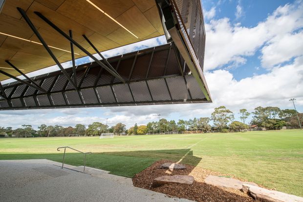 Exterior view from beneath a contemporary covered pavilion or sports facility, looking across a large grassy playing field. The structure features a timber-lined soffit, exposed black steel framing, and perforated metal screening along the cantilevered edge. A paved access area with handrails and landscaped edging occupies the foreground, with open sky and trees beyond.