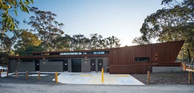 Wide exterior view of a contemporary, low-rise building in a wooded setting. The facade combines dark masonry, large dark garage or service doors, narrow clerestory glazing, and a prominent rust-brown vertically ribbed metal-clad volume with an angled roofline. A paved forecourt with bollards occupies the foreground.