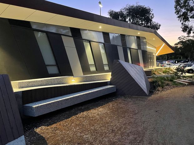 Exterior view of a contemporary low-rise building at dusk, featuring a sharply angled roof canopy, dark and light patterned facade panels, tall narrow windows, concealed linear lighting, and sculptural built-in concrete seating along the landscaped frontage. A parking area and trees are visible in the background.