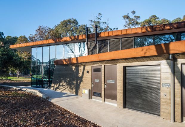Exterior view of a contemporary low-profile building in a landscaped setting. The facade combines extensive floor-to-ceiling glazing, horizontal orange-brown metal cladding and projecting bands, dark louvred screening, and a concrete-toned lower level with service doors and a roller shutter. The image appears to show a commercial or community facility rather than a residence.