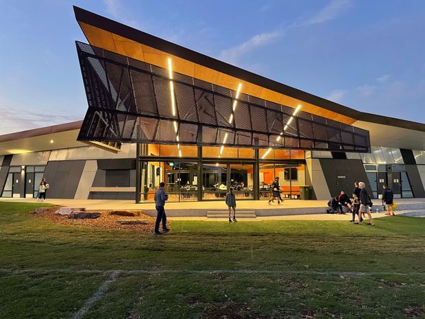 A modern public or commercial building photographed from the landscaped forecourt at dusk. The facade features angular forms, dark grey cladding, extensive glazing, warm timber-toned soffits, and a prominent projecting black perforated-metal canopy with integrated linear lighting. People gather near the entrance and visible interior activity suggests a public venue or hospitality space.