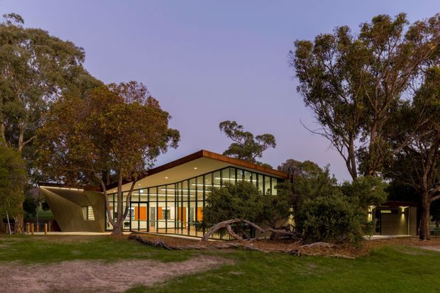 Single-storey contemporary pavilion-like building set among mature trees, photographed at dusk. The structure has extensive floor-to-ceiling glazing with dark slender frames, a low gently pitched roof with timber-toned soffit or fascia, and a distinctive projecting angled entrance or canopy volume. Interior lighting glows warmly through the glass, while landscaped lawn and fallen branches surround the building.