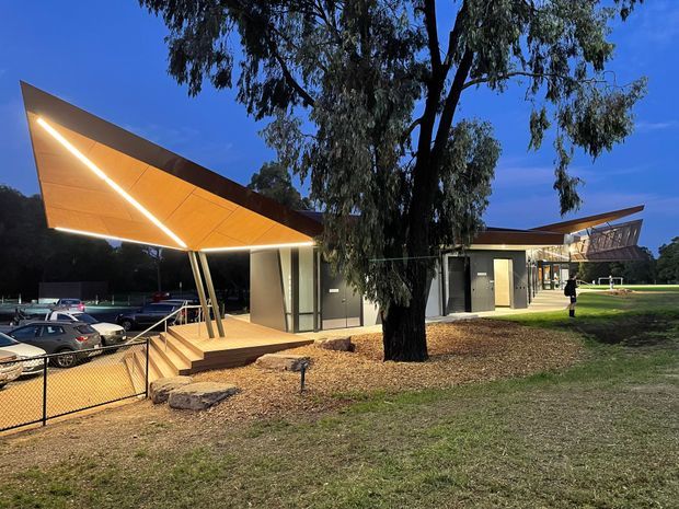 Twilight exterior view of a contemporary low-rise building with sharply angled projecting roof canopies, warm linear integrated lighting, glazed walls, and slender structural supports. The building sits in a landscaped grassy setting with mature eucalyptus trees, exposed aggregate or gravel areas, steps, and a chain-link boundary fence. Parked cars are visible beside the building.