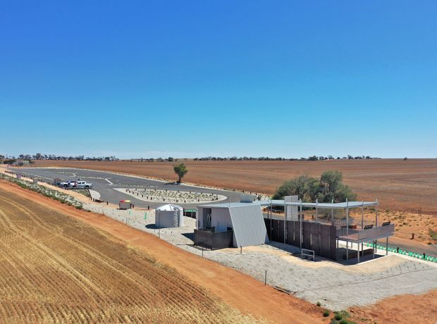 Aerial view of a modern rural property set within expansive cultivated farmland. The low, angular building has corrugated metal cladding, flat roof forms, open covered outdoor areas, and a broad concrete-paved forecourt. A curved driveway and landscaped entrance sit beside the contemporary structure, with dry agricultural fields extending to the horizon.