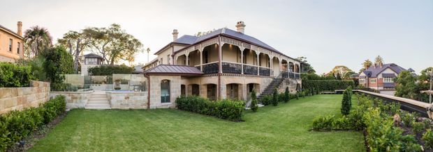 Panoramic exterior view of a substantial two-storey heritage-style residence with a hipped metal roof, wraparound covered veranda, ornate timber balustrades and posts, rendered masonry upper walls, and stone-clad lower walls. The home is set within a formal landscaped garden with expansive lawn, clipped hedges, terraces, and neighboring traditional residences visible beyond.