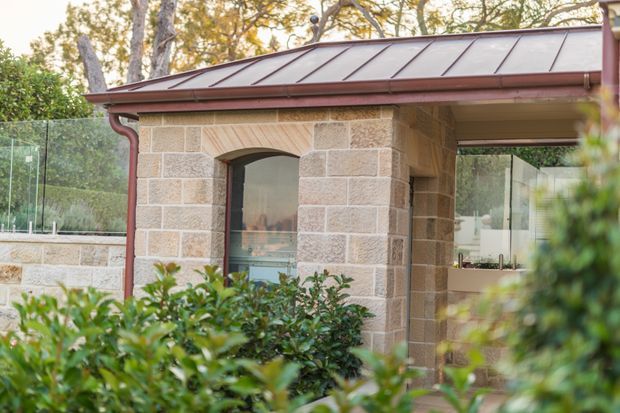 Exterior view of a compact stone-clad building or pavilion, featuring pale rendered masonry blocks, an arched window opening, large frameless glass panels, and a dark standing-seam metal roof with matching reddish-brown gutters. Dense shrubs partially screen the foreground, while mature trees are visible behind the structure.