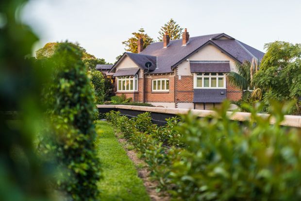 Exterior view of a traditional brick suburban house, partially framed by lush garden vegetation. The two-storey dwelling has a pitched tiled roof with multiple brick chimneys, gabled roof sections, cream rendered upper walls, dark timber weatherboard detailing, and white-framed windows. A landscaped garden path and planting occupy the foreground.