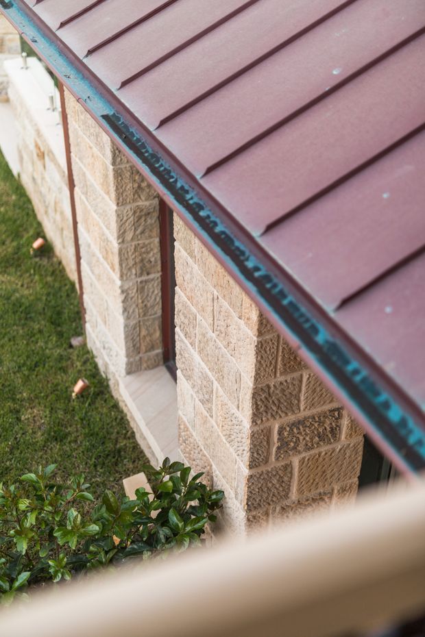 Close-up exterior architectural detail showing a maroon metal pitched roof with raised standing seams and matching metal edging above textured tan masonry block walls. A narrow paved path and landscaped planting are visible beside the building.