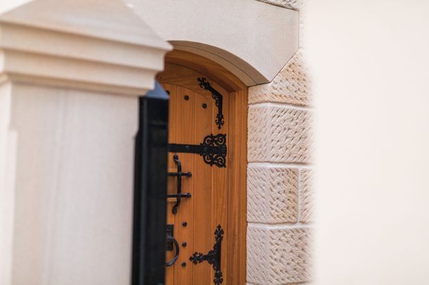 Close-up exterior view of an ornate arched timber entry door with dark wrought-iron strap hinges, handles, and decorative fittings. The doorway is framed by pale textured masonry with carved horizontal blocks and a projecting classical-style column or pilaster in the foreground.