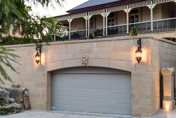 Exterior view of a substantial heritage-style residence with a lower-level double garage integrated into a textured masonry retaining wall. The upper level features a covered verandah with ornate dark metal balustrades, decorative timber posts and fretwork, a hipped tiled roof, and traditional wall-mounted lanterns. A landscaped rocky edge and mature greenery frame the property.