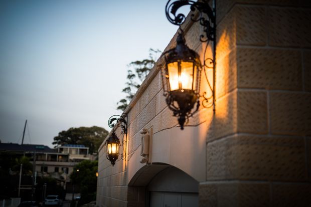 Exterior architectural detail photographed at dusk, showing a pale textured masonry wall with an arched garage opening and ornate black wrought-iron wall lanterns casting warm light. A residential or institutional building and trees are visible in the background, but the illuminated facade and lighting fixtures dominate the frame.