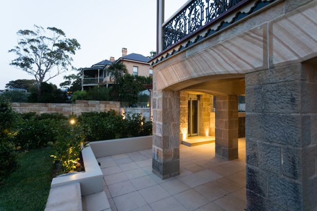 Exterior architectural view of a substantial stone-clad residence, focused on a covered terrace or entry arcade with broad square columns, a vaulted stone soffit, tiled paving, and ornate dark metal balustrading above. Landscaped hedges and a neighbouring traditional home are visible beyond the terrace. Warm interior lighting glows through the opening, creating a refined dusk atmosphere.