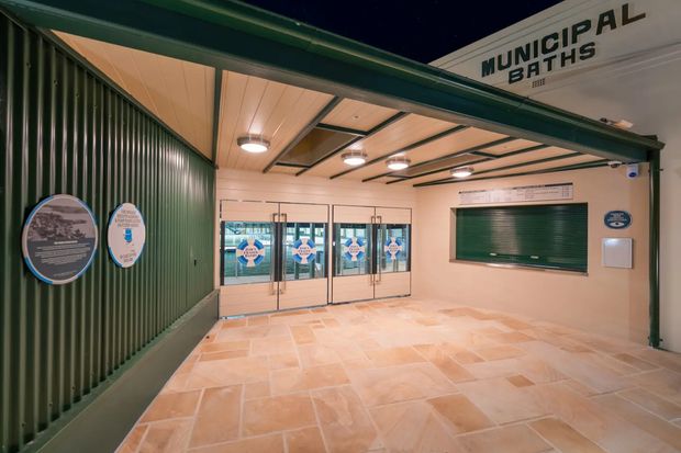 Covered public entrance or forecourt to municipal baths, with a dark green corrugated metal wall, cream rendered wall surfaces, tiled paving, glazed double doors with informational graphics, exposed dark structural beams, and illuminated signage reading “Municipal Baths.”