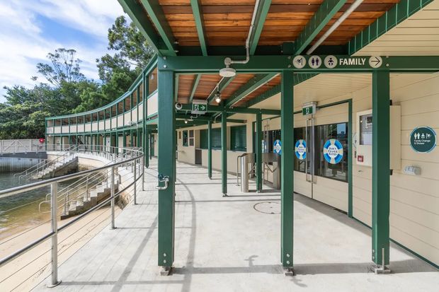 Exterior covered walkway and entrance to a public aquatic or leisure facility. The structure features green-painted steel posts and beams, exposed timber decking beneath the canopy, pale horizontal wall cladding, glazed doors, stainless-steel accessibility railings, and clear family amenity signage. A curved building wing and water feature are visible to the left, with landscaped trees beyond.