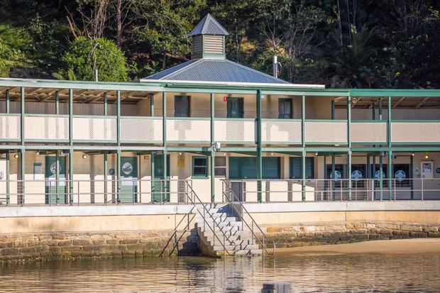 A two-storey waterfront building with a long covered verandah, pale rendered walls, green-painted metal posts and railings, patterned privacy screens, and a central pyramid-roofed cupola. The lower level opens toward the water via a broad concrete terrace and central staircase, while dense trees form the background.