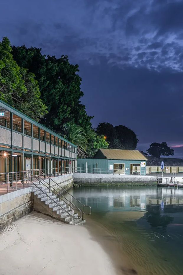 A dusk exterior view of a waterside recreational or commercial facility beside a calm enclosed body of water. The scene features a long two-storey glazed building with exposed timber roof framing and metal mesh or panel balustrades, a smaller green-clad gabled utility building, concrete retaining edges, and a broad stair descending into the water. Dense tropical vegetation surrounds the site beneath a dramatic dark blue sky.