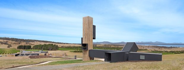 Wide exterior view of a contemporary, low-profile dark-clad building in an open rural landscape. The structure features a prominent tall vertical timber-clad tower, flat and pitched roof forms, and a long horizontal wing. Agricultural fields, distant hills, and a body of water are visible in the background.