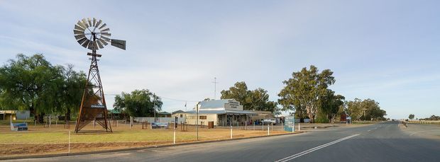 Wide exterior streetscape showing a rural or regional roadside property with a prominent traditional windmill on a tall metal tower, low single-storey buildings, fencing, mature trees and an expansive paved road under an open sky.