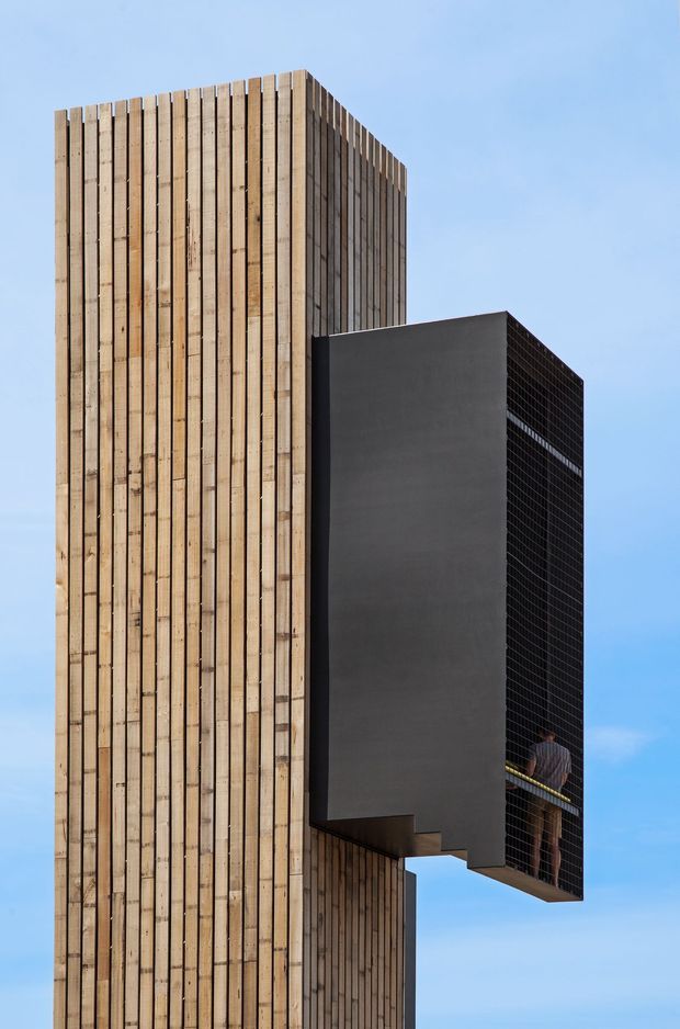 Exterior architectural photograph of a tall, highly sculptural building against a clear blue sky. The main volume is clad in closely spaced vertical timber boards, with a projecting dark charcoal rectangular box featuring a smooth solid side wall and a gridded dark facade. A person is visible within the projecting volume, emphasizing its scale and cantilevered form.