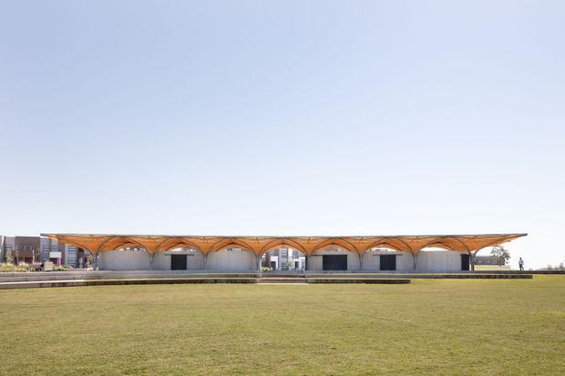 Wide exterior view of a low, elongated contemporary building set behind an expansive lawn. The structure features a distinctive undulating canopy roof with repeated arched bays, slender supports, and glazed or open frontage. Additional modern buildings are visible in the background under a clear sky.