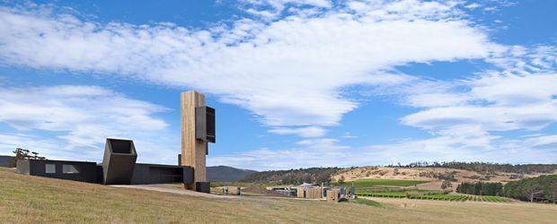 A low, angular dark-clad building set in a broad rural landscape, with a prominent tall rectangular timber-clad vertical element and sculptural projecting volume. Rolling cultivated fields and wooded hills extend behind it beneath a partly cloudy blue sky.