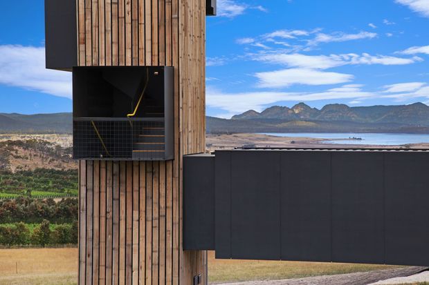 Exterior architectural photograph of a contemporary building set against a broad rural valley, lake, and mountain backdrop. The facade combines vertically arranged timber cladding with dark charcoal wall panels and a projecting, recessed stair or balcony volume with black tiled surfaces and a bright yellow handrail. The composition emphasises the building's strong vertical and horizontal geometric forms, with clear blue sky and expansive landscape beyond.