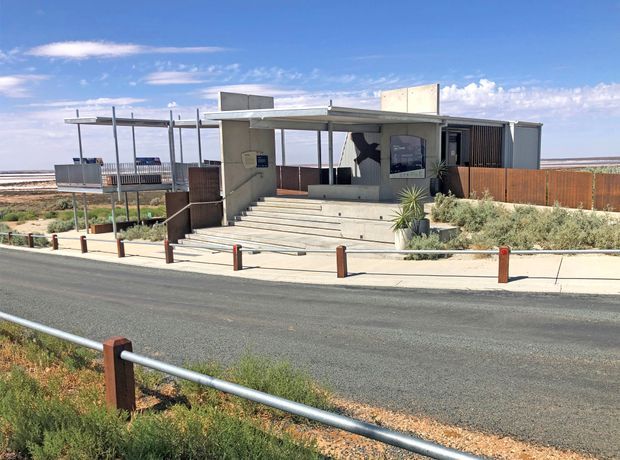 A low, contemporary pavilion-like building beside a broad sandy coastal or tidal landscape. The structure has a flat roof, exposed concrete entrance steps and walls, extensive dark-framed glazing, vertical metal screening, timber privacy fencing, and sparse drought-tolerant landscaping. A paved road and simple metal-and-timber pedestrian barriers run across the foreground.