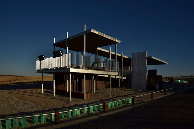 Exterior view of a contemporary elevated, flat-roofed building on a very open, sparsely landscaped site. The structure features slender posts, exposed upper-level decks with metal balustrades, dark recessed glazing, vertical timber-like screening, and a prominent concrete or grey panel wall. Strong low-angle sunlight creates deep shadows against a clear dark blue sky.