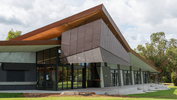 Contemporary single-storey building exterior with a dramatic asymmetric sloping roof and projecting central upper volume clad in perforated metal screens. The facade combines dark metal panels, extensive floor-to-ceiling glazing, and warm rust-coloured roof soffits, set within landscaped grounds under a partly cloudy sky.