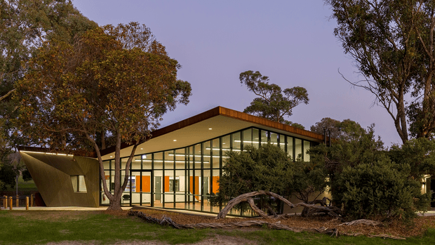 A low, modern pavilion-like building set within a mature eucalyptus landscape at dusk. The structure features a broad shallow-pitched roof with deep timber-toned fascia, extensive floor-to-ceiling glazing in a dark slender frame, and warm interior lighting visible through the glass. A sculptural angular solid volume projects from the glazed facade, while fallen branches, shrubs, lawn, and established trees frame the building.