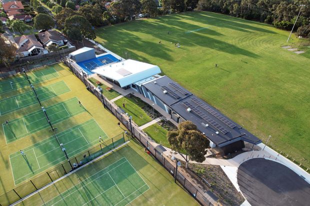 Aerial view of a large contemporary community or school sports facility beside expansive playing fields. Multiple fenced green tennis courts dominate the foreground, with a blue swimming pool, low modern buildings featuring extensive rooftop solar panels, paved access areas, and landscaped suburban surroundings.
