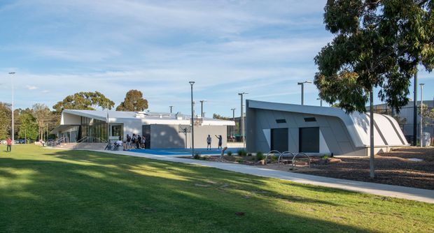 Wide exterior view of a contemporary low-rise civic or commercial building complex set within landscaped grounds. The buildings feature angular rooflines, smooth grey cladding, extensive glazing, and a distinctive curved-roof volume, with paved pedestrian paths, lawn, mature trees, and outdoor bicycle racks visible.