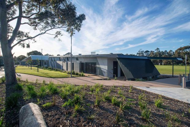 Low, single-storey contemporary building set within landscaped grounds, featuring a dark ribbed metal-clad projecting entry volume, light grey walls, expansive glazing, and a shallow low-pitched roof. A paved pedestrian approach, lawn, mature trees, and open recreational grounds surround the building.