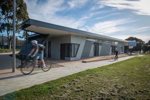 Low, single-storey contemporary building with an angular grey-clad facade, broad projecting roof canopy, extensive glazing, and a paved pedestrian path alongside a grassy open area. Cyclists are passing in the foreground and beside the building, suggesting a public or recreational facility.