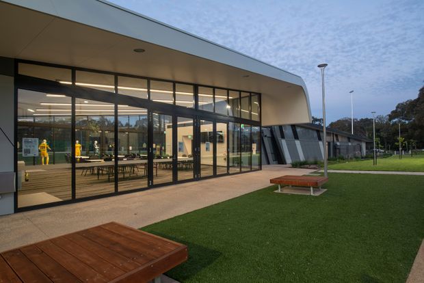 Exterior view of a contemporary public or commercial building with a broad curved roof canopy, extensive floor-to-ceiling glass curtain walls, dark slim frames, and a landscaped lawn. Interior tables and display elements are visible through the glazing, while timber benches and paved paths occupy the foreground.