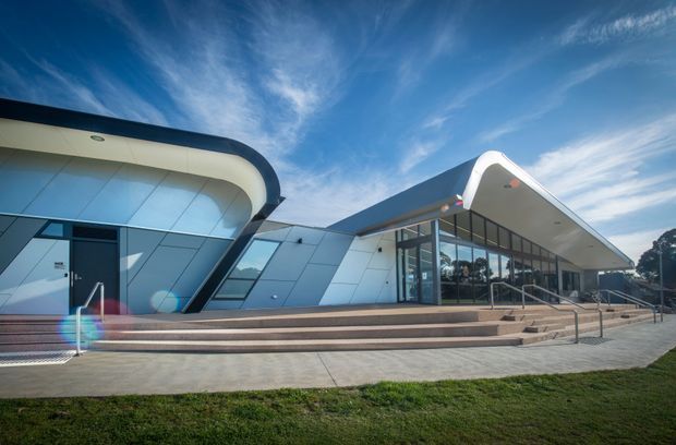Exterior view of a contemporary public or commercial building with dramatic curved roof forms, angular silver-grey wall panels, extensive glazed frontage, broad entry steps, and accessible handrails. The building sits in a landscaped open setting beneath a bright blue sky.