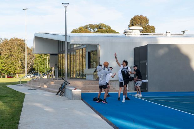 Exterior view of a contemporary low-rise sports or recreation building beside a blue outdoor court. The building has a sharply projecting flat-roofed canopy, extensive floor-to-ceiling glazing, corrugated light-metal cladding, dark grey wall panels, rooftop mechanical vents, and concrete steps and paving. Several people are playing basketball on the court, with landscaped trees and lawn surrounding the facility.