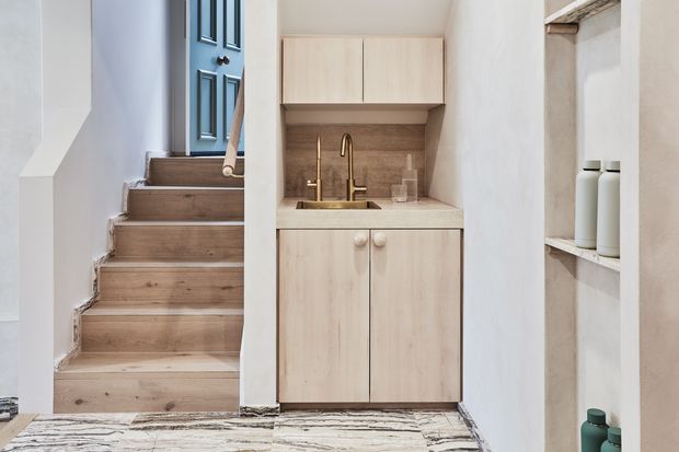 Compact light-wood kitchenette or beverage station built beneath an upper cabinet, with a small sink, brass-toned tapware, and pale wood cabinetry. A timber staircase with a blue paneled door rises alongside it, while open shelving displays minimal ceramic vessels. The space has a warm, refined, contemporary feel with natural stone-patterned flooring.