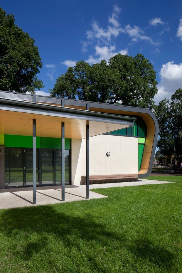Exterior view of a distinctly contemporary low-rise building with a curved projecting roof and fascia, slim dark structural posts, extensive dark-framed glazing, pale masonry walling, and vivid green accent panels. The building sits in a manicured lawn beneath mature trees and a bright blue sky.