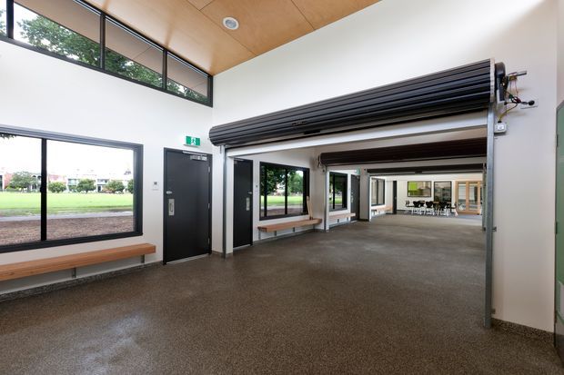 A bright, modern open-plan commercial interior with polished concrete flooring, white walls, a warm timber-lined ceiling, extensive black-framed glazing, and exposed roller doors. Built-in timber benches line the windows, with a further open room and meeting area visible beyond. The space appears to be a flexible community, office, or commercial facility with a clean industrial-modern character.