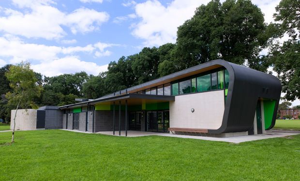 A low-rise contemporary building set within a landscaped lawn and mature trees. The design features a long, gently sloping roofline, extensive green-tinted glazing, dark ribbed curved cladding wrapping the right-hand volume, pale rendered wall panels, and bright green accent reveals and interior elements. A covered entrance walkway runs along the front facade.
